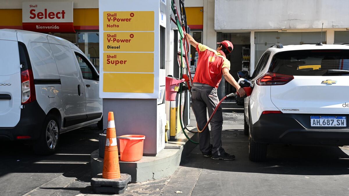 Un empleado de Shell, vestido con uniforme rojo y amarillo, carga combustible en un SUV blanco en una estación de servicio Shell con un cartel de precios