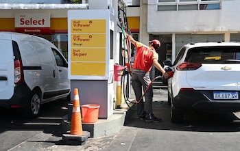 Un empleado de Shell, vestido con uniforme rojo y amarillo, carga combustible en un SUV blanco en una estación de servicio Shell con un cartel de precios
