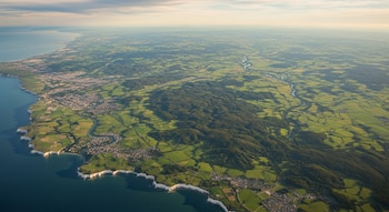 Imagen aérea del mar, acantilados blancos, campos verdes divididos, bosques, un río sinuoso y asentamientos costeros bajo un cielo despejado.
