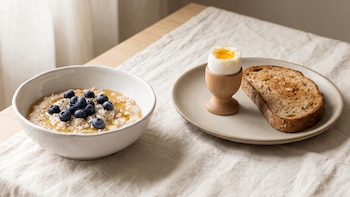Tazón blanco de avena con arándanos junto a un plato con huevo pasado por agua y tostada sobre un mantel claro.