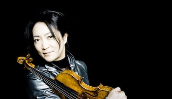 Retrato de la violinista Akiko Suwanai, una mujer de cabello oscuro con una chaqueta brillante, sosteniendo un violín de madera contra un fondo negro