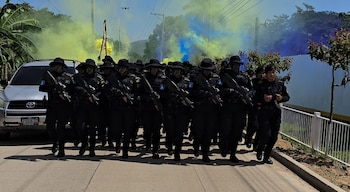 Grupo de agentes uniformados de negro con sombreros y fusiles, caminando en una calle. Humo amarillo y azul flota en el aire. Un vehículo utilitario compacto plateado a la izquierda