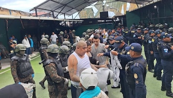 Agentes de policía y soldados armados con cascos y chalecos rodean a hombres de civil en una zona exterior con suelo verde y techo metálico