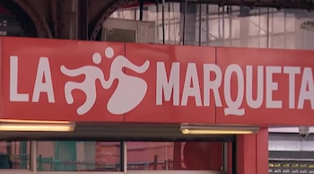 Primer plano de un letrero rectangular rojo con la frase "LA MARQUETA" en letras blancas grandes y un logo de dos figuras estilizadas