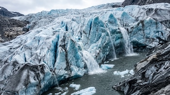 Un glaciar de hielo azul y blanco con grietas, del cual caen cascadas de agua de deshielo a un lago turbio con bloques de hielo flotando.