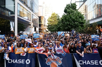 Una multitud diversa de personas vestidas con ropa de los New York Knicks se alinea detrás de una valla con el logo de Chase, sosteniendo carteles en una calle de la ciudad