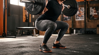 Persona vista desde las piernas hasta los hombros realizando una sentadilla con barra y discos en un gimnasio.