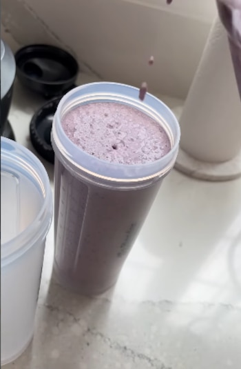 Un vaso de plástico transparente lleno con un batido morado espumoso sobre una encimera clara de cocina