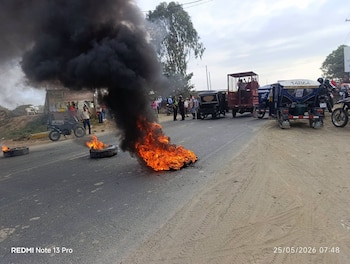 Carretera bloqueada con dos neumáticos en llamas y denso humo negro. Varias personas y vehículos como mototaxis y motocicletas están detenidos cerca