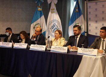 Seis personas, cinco hombres y una mujer, sentadas en una mesa con micrófonos y tarjetas de identificación. Detrás hay tres banderas y un logotipo institucional