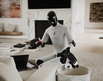 Robot humanoide Figure AI blanco y negro agachado en un salón. Sostiene un coche rojo y deposita una pelota en una cesta. Mesa de centro, sofá y portátil visibles