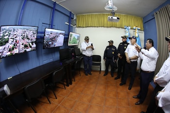 Sala con tres oficiales uniformados, tres hombres de civil, tres pantallas con imágenes urbanas y mapa, escritorios con computadoras y proyector