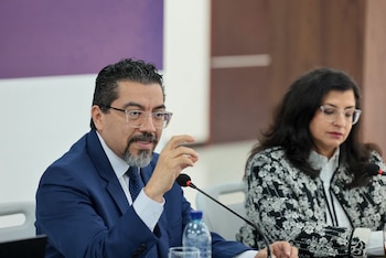 Un hombre con traje azul y gafas habla ante un micrófono en una mesa junto a una mujer con gafas y chaqueta estampada