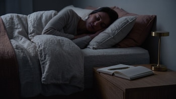 Una mujer acostada en una cama junto a una mesa de noche de madera con un cuaderno abierto, un bolígrafo y una lámpara
