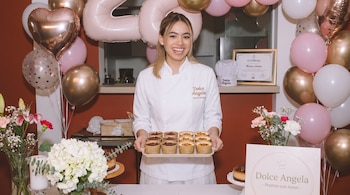 Mujer chef sonriente con uniforme blanco sostiene bandeja con doce postres individuales. Alrededor hay globos, flores, pasteles y un certificado.