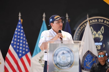 Hombre con gorra azul y camisa blanca habla en micrófono. Podio con escudo policial de Guatemala, bandera de Estados Unidos y bandera de Guatemala en el fondo