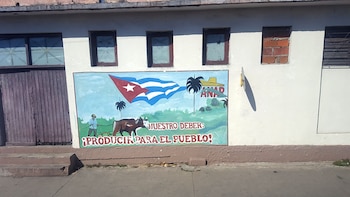 Mural en una pared blanca que muestra un hombre arando con un buey, la bandera cubana y palmeras, con texto rojo debajo