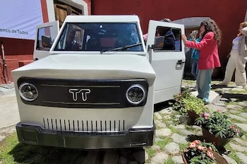 Vista frontal y lateral de un auto eléctrico blanco compacto con el logotipo 'TT' en la parrilla, sus puertas delanteras están abiertas y personas lo observan