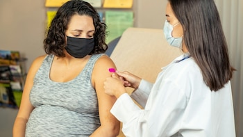Una trabajadora de salud con mascarilla coloca un parche rosado en el brazo de una mujer embarazada que viste camiseta gris y mascarilla negra