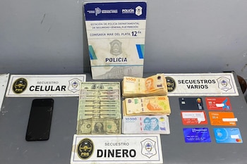 Un teléfono celular negro, billetes de dólares y pesos argentinos, varias tarjetas de crédito y carteles policiales colocados sobre una mesa