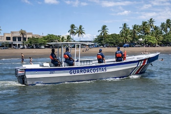 Lancha de Guardacostas con cuatro oficiales a bordo y el número 397, agua de mar, playa con arena, palmeras, varias personas en la orilla y bañistas.
