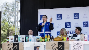 Una mujer con chaleco azul habla por micrófono ante un panel institucional, frente a una mesa con paquetes de café, frascos y botellas