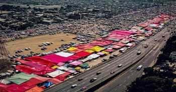 Vista aérea de un mercado extenso con filas de carpas multicolores, una autopista con vehículos, estacionamientos llenos de coches y edificios urbanos en el fondo