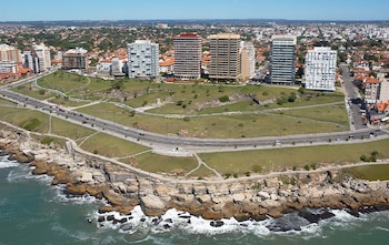 Vista aérea de una costa rocosa con olas, una avenida con vehículos, un parque con hierba y varios edificios de altura