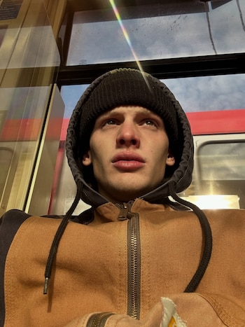 Primer plano de Freddy Brazier, un hombre joven con gorro negro y chaqueta marrón con capucha, mirando hacia arriba. Ventanas y reflejos de luz solar de fondo