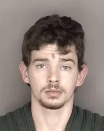 Hombre joven con pelo castaño rizado, ojos claros, barba incipiente y una marca en la frente, vistiendo ropa oscura sobre un fondo claro