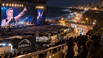 Escenario con pantallas gigantes, multitud de asistentes en un concierto nocturno, vía iluminada con vehículos y personas en un puente elevador
