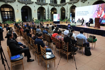 Personas sentadas en filas de sillas ante un escenario con panelistas, una pantalla informativa y la bandera de Guatemala
