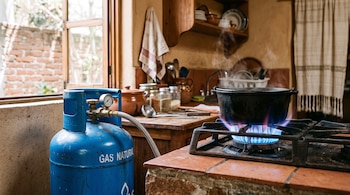 Un cilindro metálico azul conectado por una manguera a una hornilla con llama azul y una olla de hierro que emite vapor.