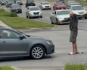 Un hombre calvo con barba, sudadera y bermudas, apunta una pistola hacia el frente de un automóvil gris en una calle con varios vehículos en circulación