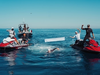 Una persona nada en mar abierto entre dos motos acuáticas rojas y una lancha con bandera mexicana bajo un cielo despejado