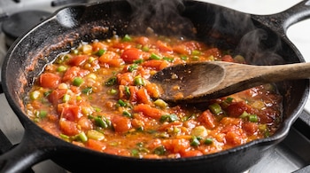 Sartén de hierro fundido con tomate y cebolla picados botando humo y una cuchara de madera apoyada en la mezcla.