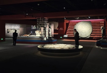 Interior de sala de exposición con techo oscuro, paredes rojas y oscuras, escaleras iluminadas, dos hombres, piedra circular tallada, estatuas, maquetas y vitrinas