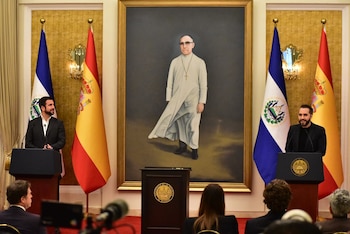 Dos hombres ante podios en una sala con banderas de El Salvador y España, con una pintura de un sacerdote al centro