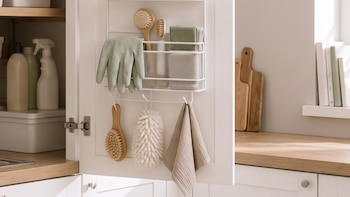 Gabinetes blancos con encimera de madera, fregadero de acero inoxidable. Armario abierto con estantes, cestas, botellas, guantes, cepillos y toallas. Ventana.