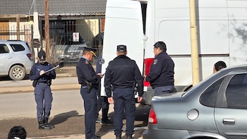 Cuatro agentes de policía en uniforme; una oficial escribe en un portapapeles; tres oficiales masculinos junto a una camioneta blanca; un automóvil gris y otro plateado