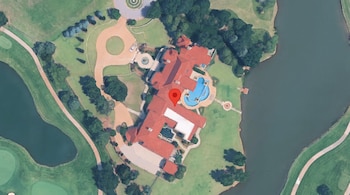 Vista satelital de una mansión con techos rojos, piscina y jardines, señalada por un marcador rojo entre un lago y áreas verdes.