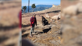 Una mujer con gafas de sol, camisa roja, pantalón caqui y mochila azul posa en un sendero montañoso con rocas, árboles secos y montañas al fondo
