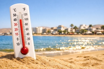 Primer plano de un termómetro blanco en la arena de una playa, marcando alta temperatura. Al fondo, una ciudad costera con edificios y palmeras, y el mar brillando bajo un cielo azul.