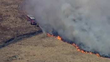 Un camión de bomberos rojo y un bombero rociando agua sobre una línea de fuego y humo denso en un campo de pasto seco, visto desde arriba