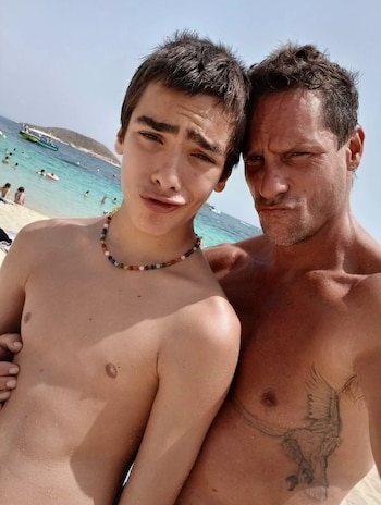 Primer plano de dos hombres, uno adulto con tatuaje de águila en el pecho y uno joven con collar de cuentas. Al fondo, playa de arena, mar azul, barcos y personas