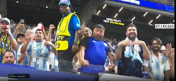 Multitud de personas en un estadio, algunos con camisetas de Argentina. Un guardia de seguridad, una pantalla con el marcador Argentina 3 Egipto 2 y un número de ayuda