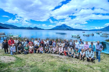 Grupo de aproximadamente cuarenta personas posando en césped, con un lago y volcanes al fondo, embarcaciones en el agua, cielo nublado