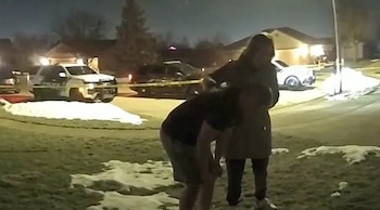 Dos personas permanecen en el césped de una zona residencial de noche frente a varias patrullas policiales y una cinta de acordonamiento