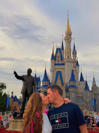 Una mujer de chaqueta rosa y un hombre con camiseta azul se besan frente a una estatua de bronce y un castillo con torres azules