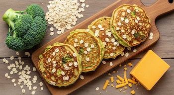 Toma cenital de cinco tortitas de brócoli y avena en una bandeja de madera, con brócoli, copos de avena y un trozo de queso cheddar al lado.